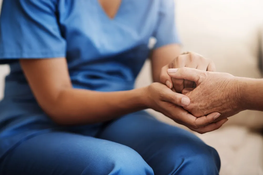 A caregiver in scrubs holding the hand of an older adult.